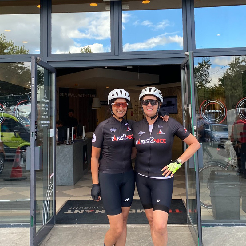 Monica Sood and Annemarie Corcoran on a training ride for ovarian cancer research Selfie of Annemarie Corcoran and Monica Sood wearing cycling helmets, sunglasses, and custom cycling jerseys during a training break in a parking area.