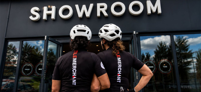 Monica Sood and Annemarie Corcoran standing side-by-side from behind, wearing branded Tile Merchant cycling jerseys and helmets, looking towards the entrance of the Tile Merchant showroom before their Paris2Nice charity cycle.