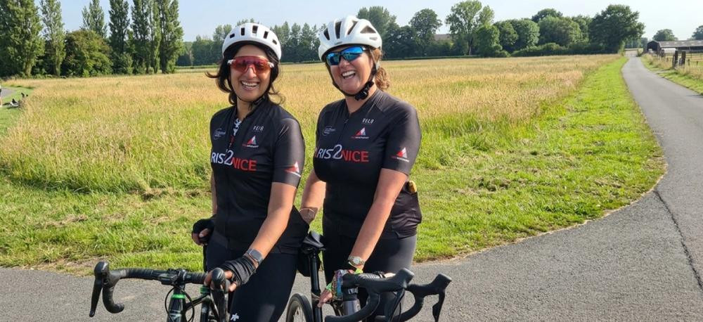 Monica and Annemarie wearing custom Tile Merchant Paris2Nice cycling jerseys Monica Sood and Annemarie Corcoran posing with road bikes on a rural path, wearing white helmets and matching black Paris2Nice cycling jerseys sponsored by Tile Merchant.