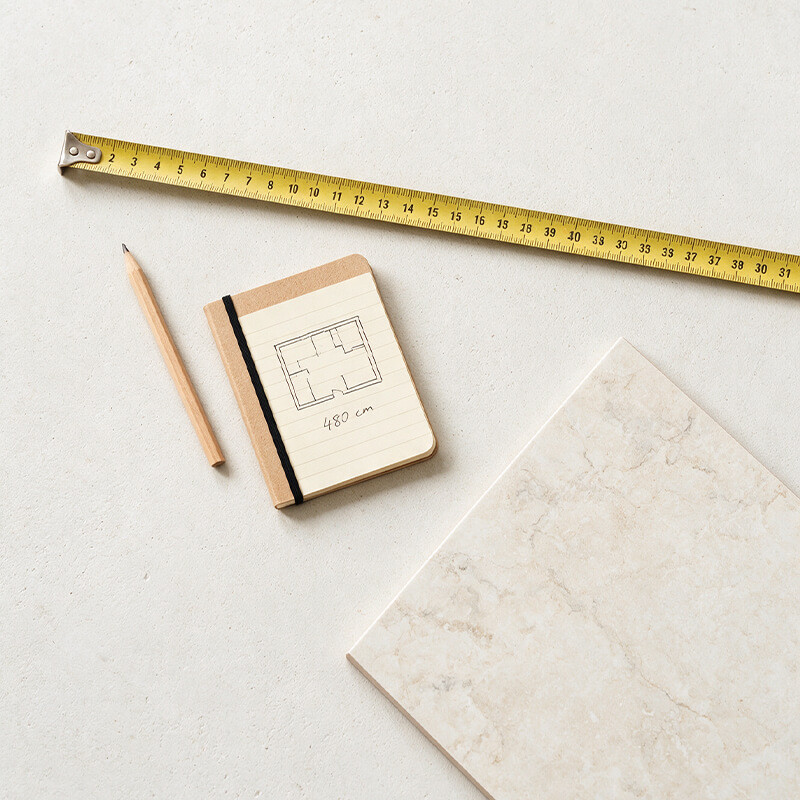 Tools for measuring a room for tiles Tape measure, pencil, notebook with a sketched floor plan and a marble-effect tile sample laid flat on a white surface