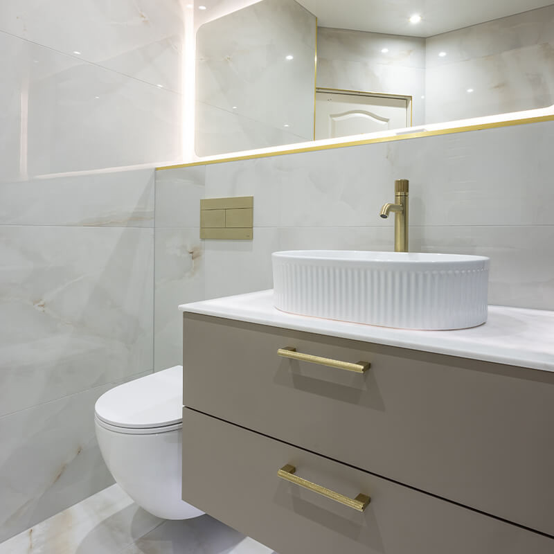 Small bathroom with large-format light marble-effect wall tiles, taupe vanity with gold handles, fluted basin and wall-hung toilet